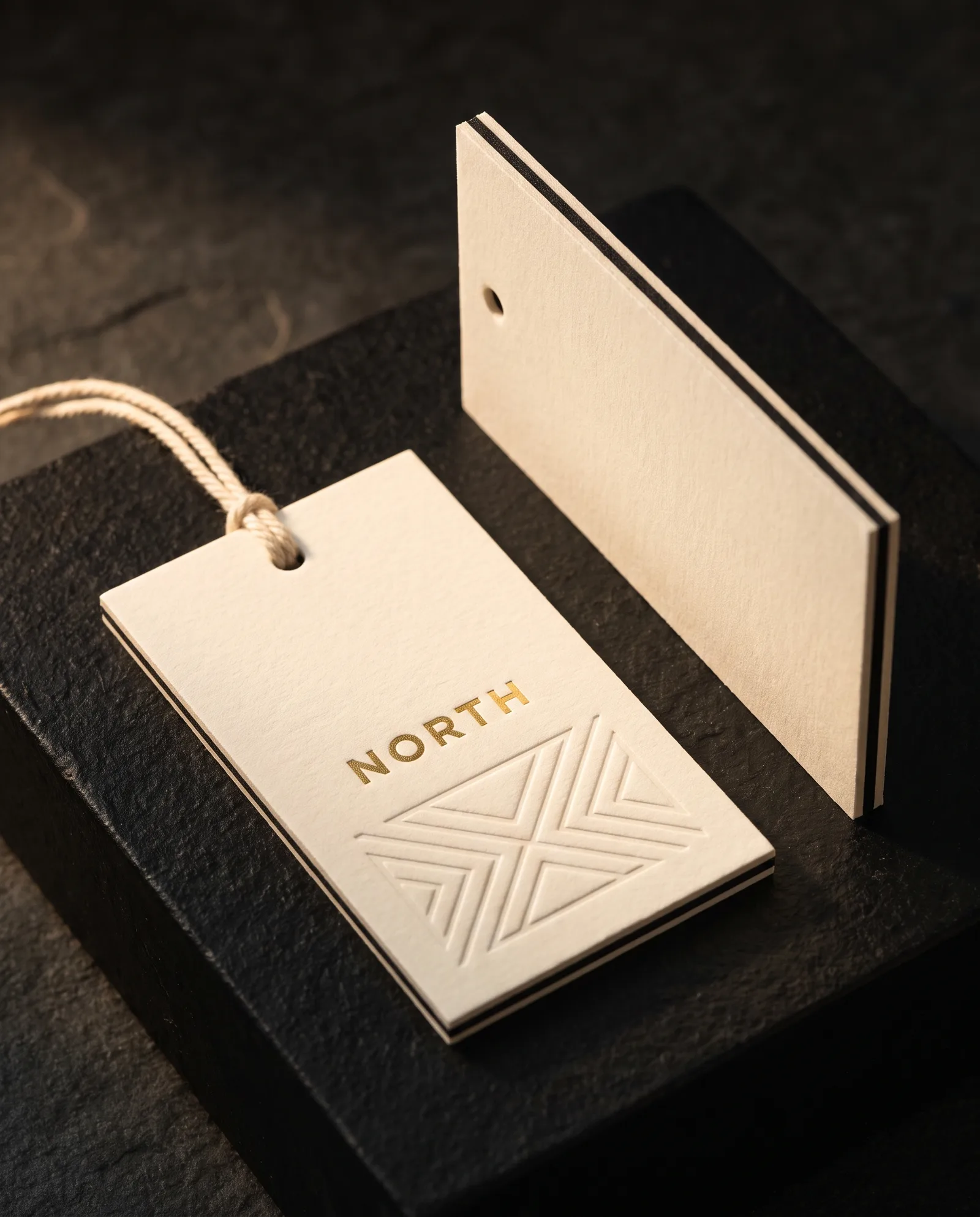Thick duplex cotton hang tag with a gold foil mark and a debossed pattern, a second tag turned to show the bonded edge.
