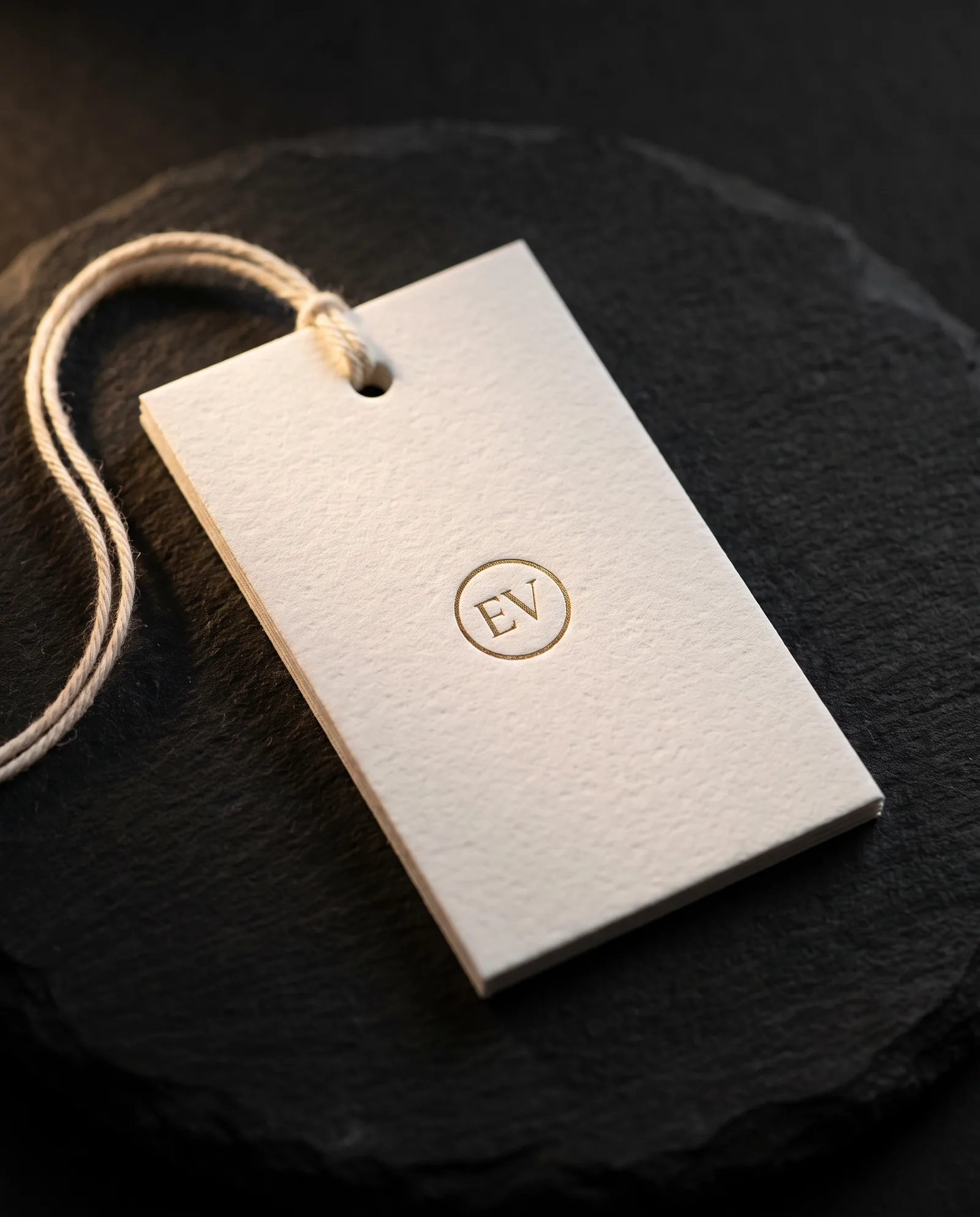 Pearl white cotton gift tag with a gold foil mark and natural cotton string, on dark stone.
