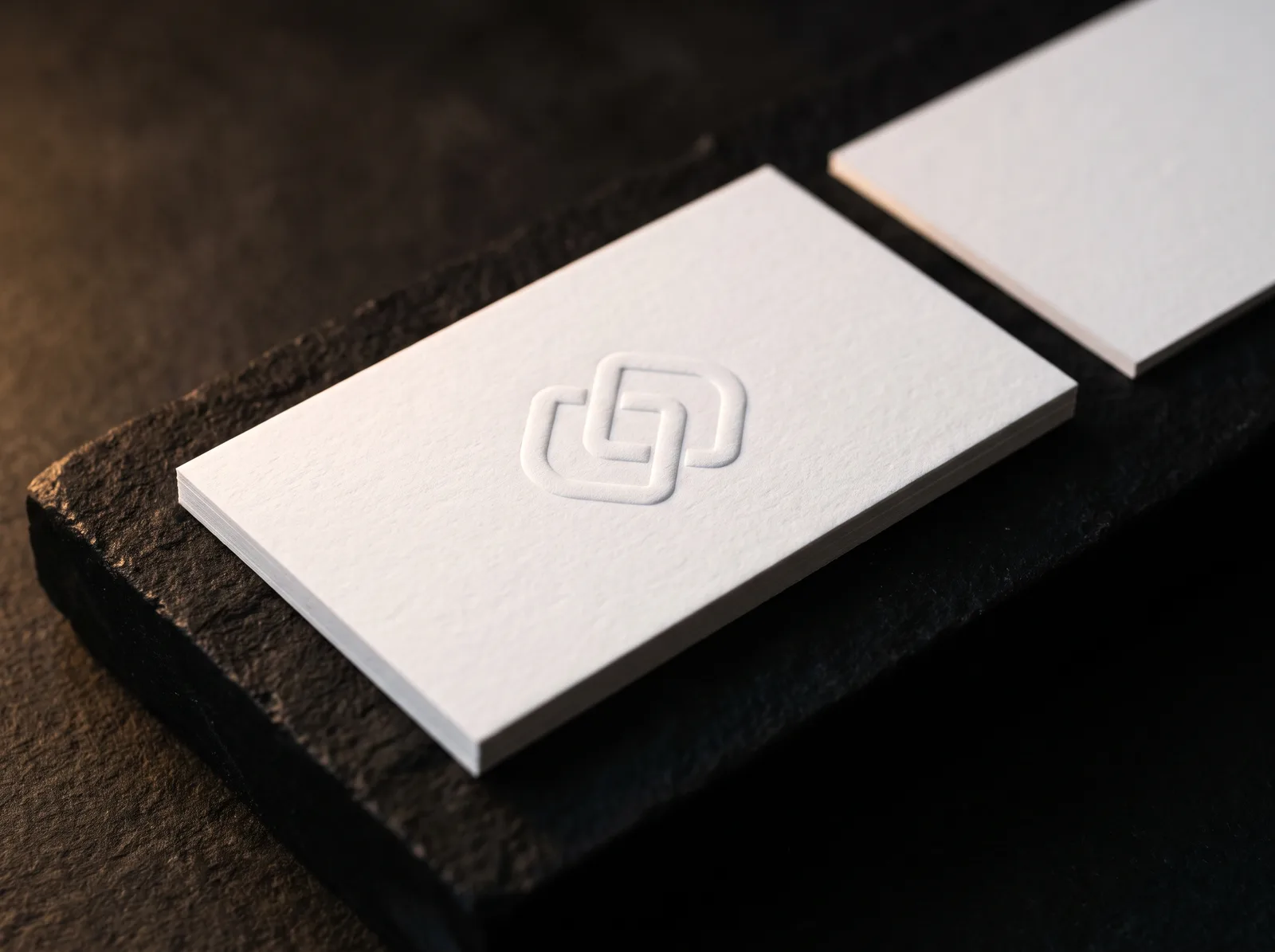 Business card with embossed logo: a blind emboss on 32pt white cotton, raised and caught by a raking light on a black stone plinth