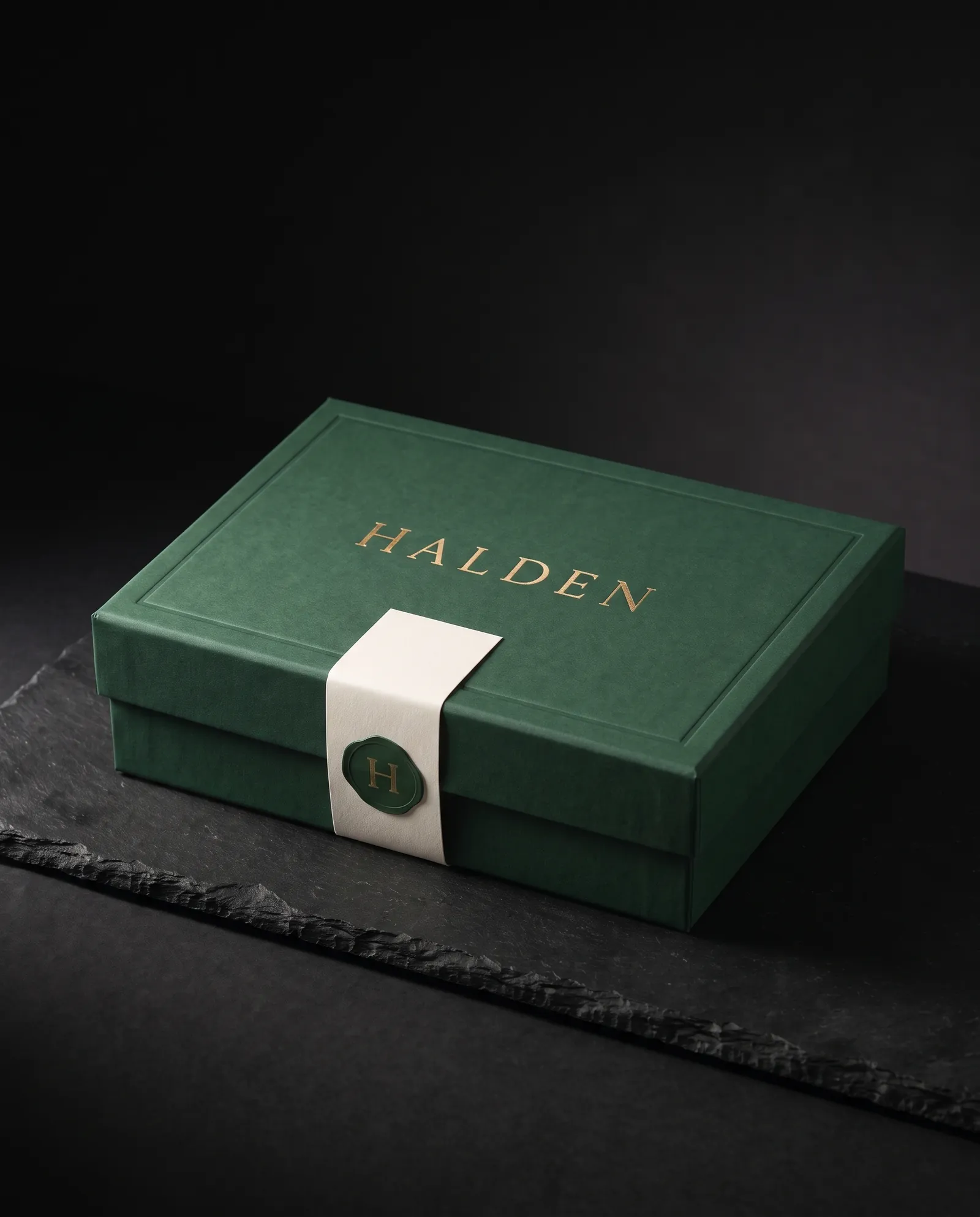 Closed forest green rigid box with a gold foil wordmark, wrapped in an ivory cotton belly band closed with a foil seal