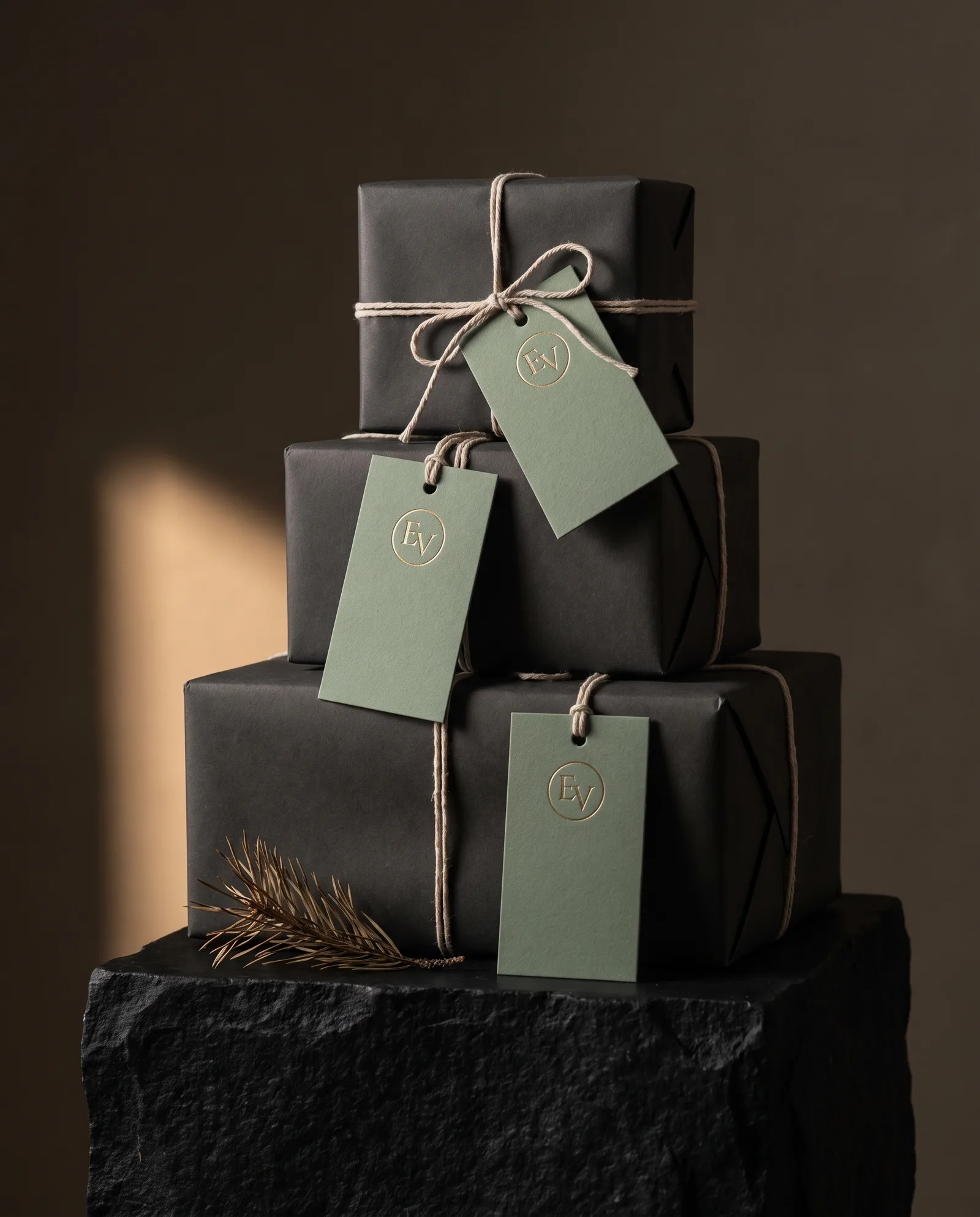 Stack of wrapped gift boxes in dark paper, each tied with cotton string and a sage cotton tag with a gold foil mark.