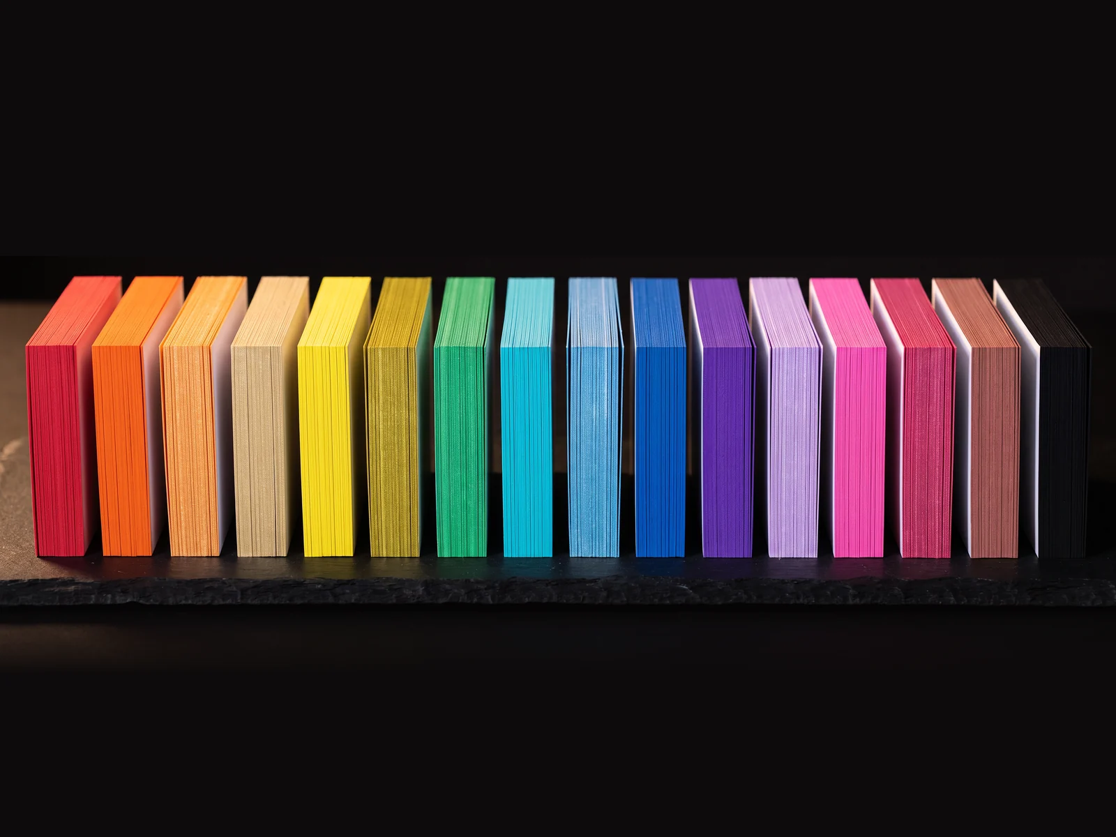 All 16 Standard painted edge colours as upright card stacks in a row, red through black, on black slate