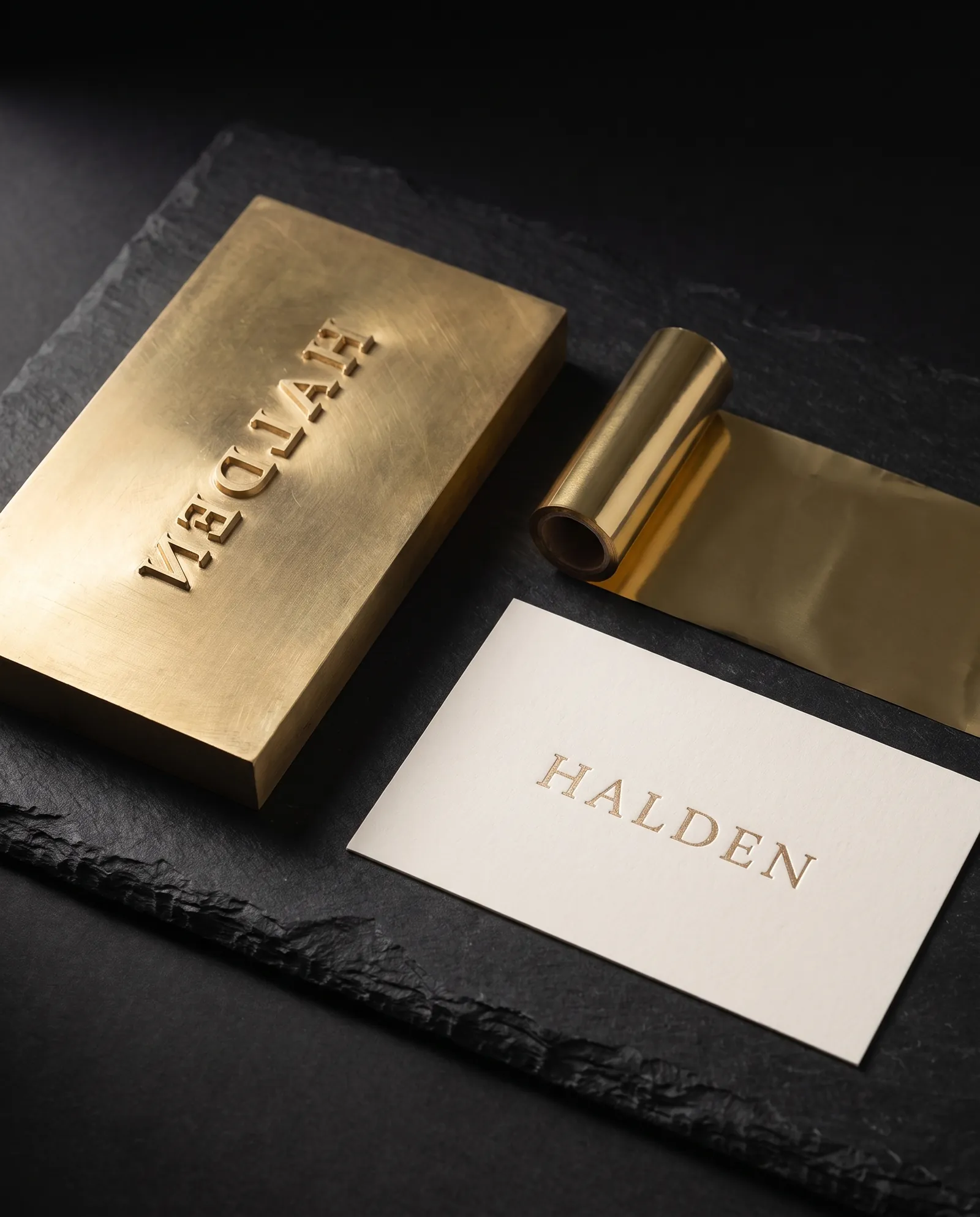 A brass foil stamping die with a mirrored wordmark, a roll of gold foil and the stamped card