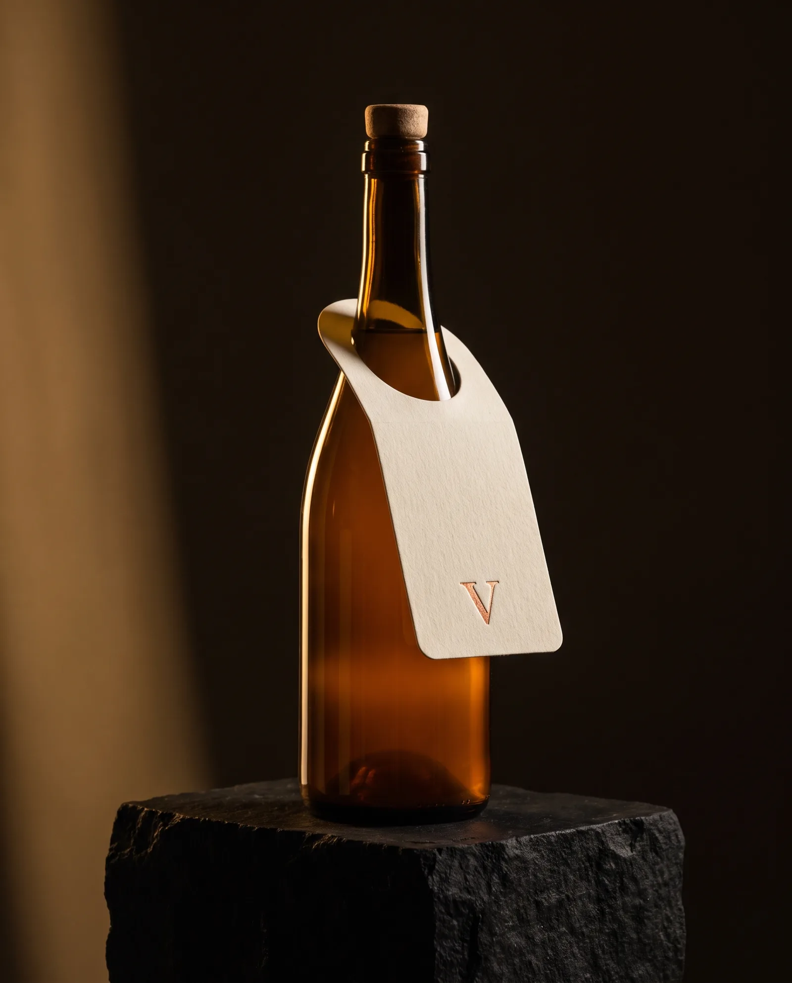 Amber glass bottle on dark stone with a die-cut cream cotton neck tag and a copper foil mark.