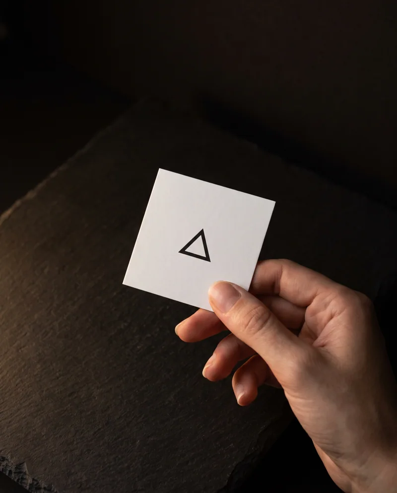 Bare hand holding a 2.5 inch square white business card over dark stone