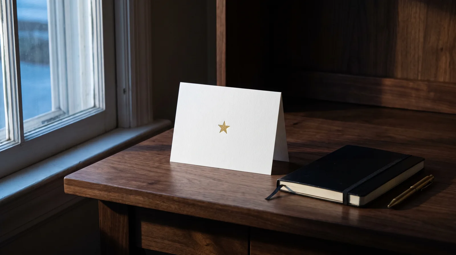 White cotton holiday card with a small gold foil star standing on a walnut desk by a window.