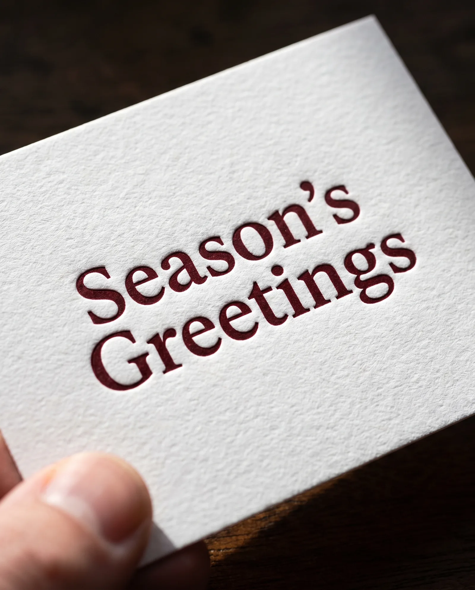 Macro of oxblood letterpress reading Season's Greetings pressed into white cotton, a thumb resting at the edge of the card.
