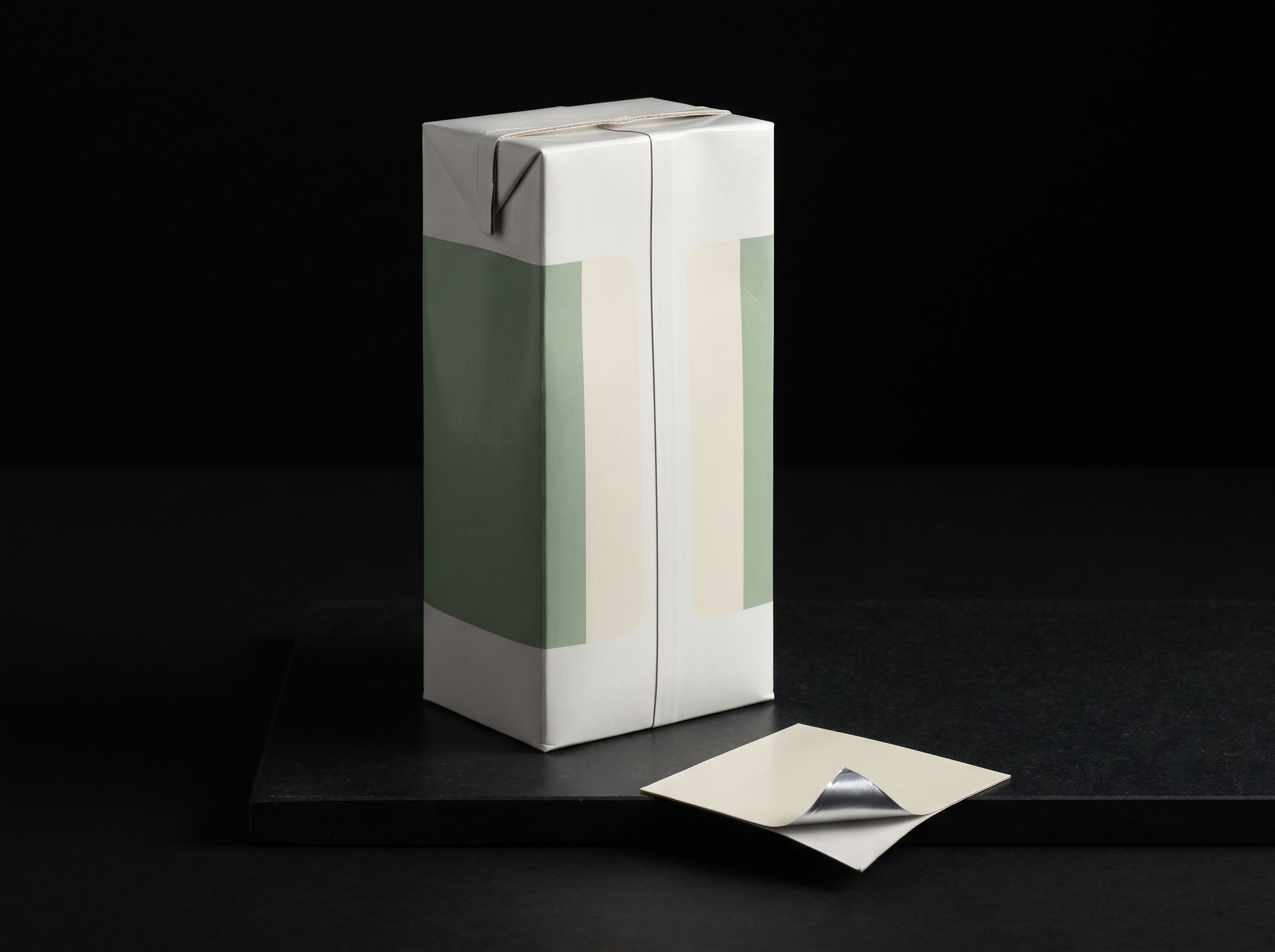 A white aseptic brick carton printed in sage-green and cream bands, with a cut piece of the laminate showing the white board and silver foil inside.