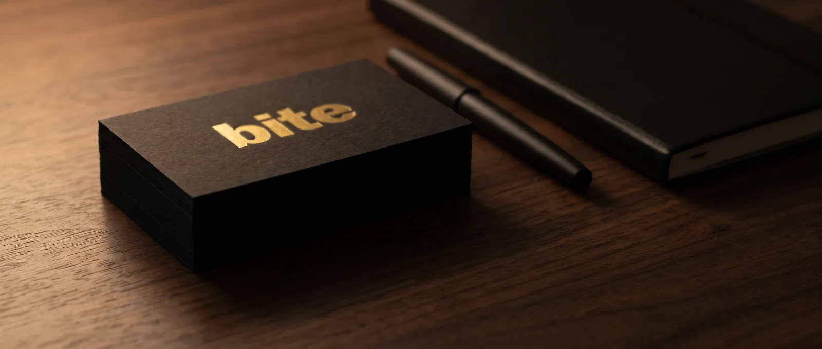 Stack of black business cards with gold foil wordmark on walnut desk beside notebook and pen