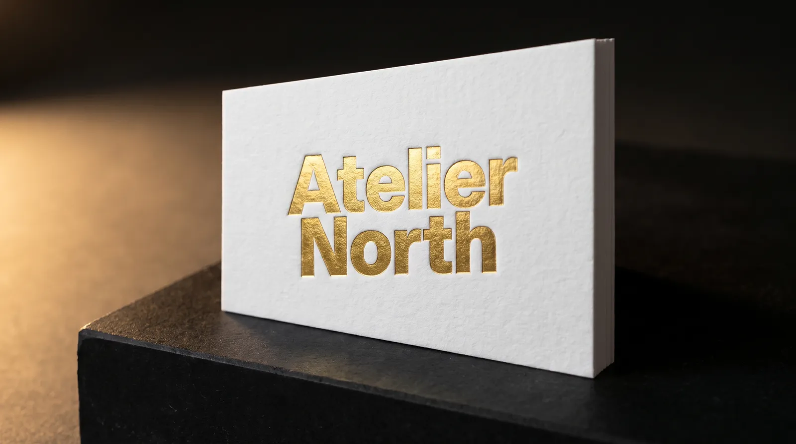 Gold foil business card on 32pt white cotton, foil wordmark catching a raking light on a black stone plinth