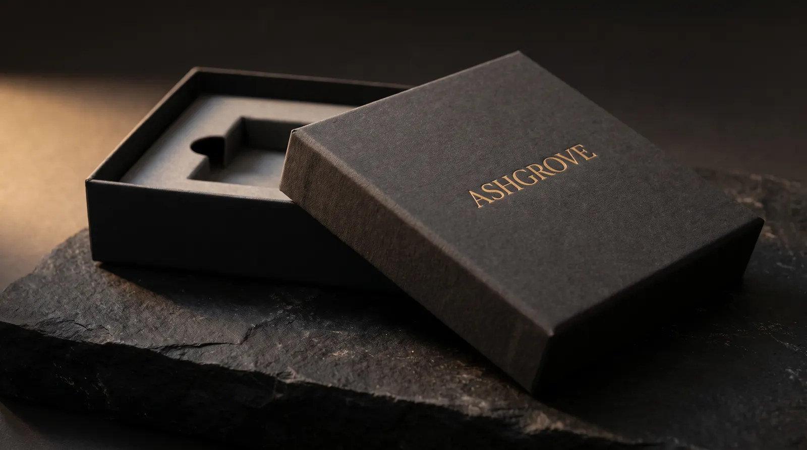 Charcoal uncoated rigid gift box with gold hot foil wordmark on lid, open on black stone