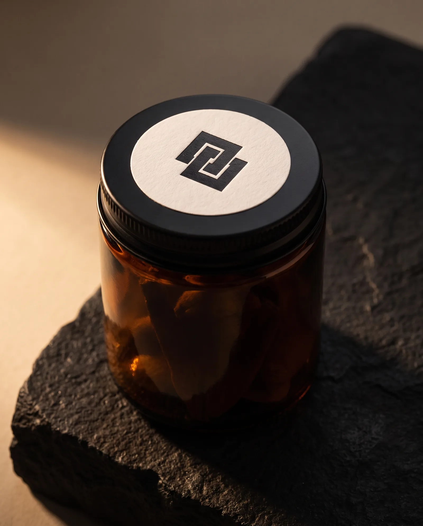 Amber jar with a black lid sealed with a round white sticker carrying a matte black foil mark.