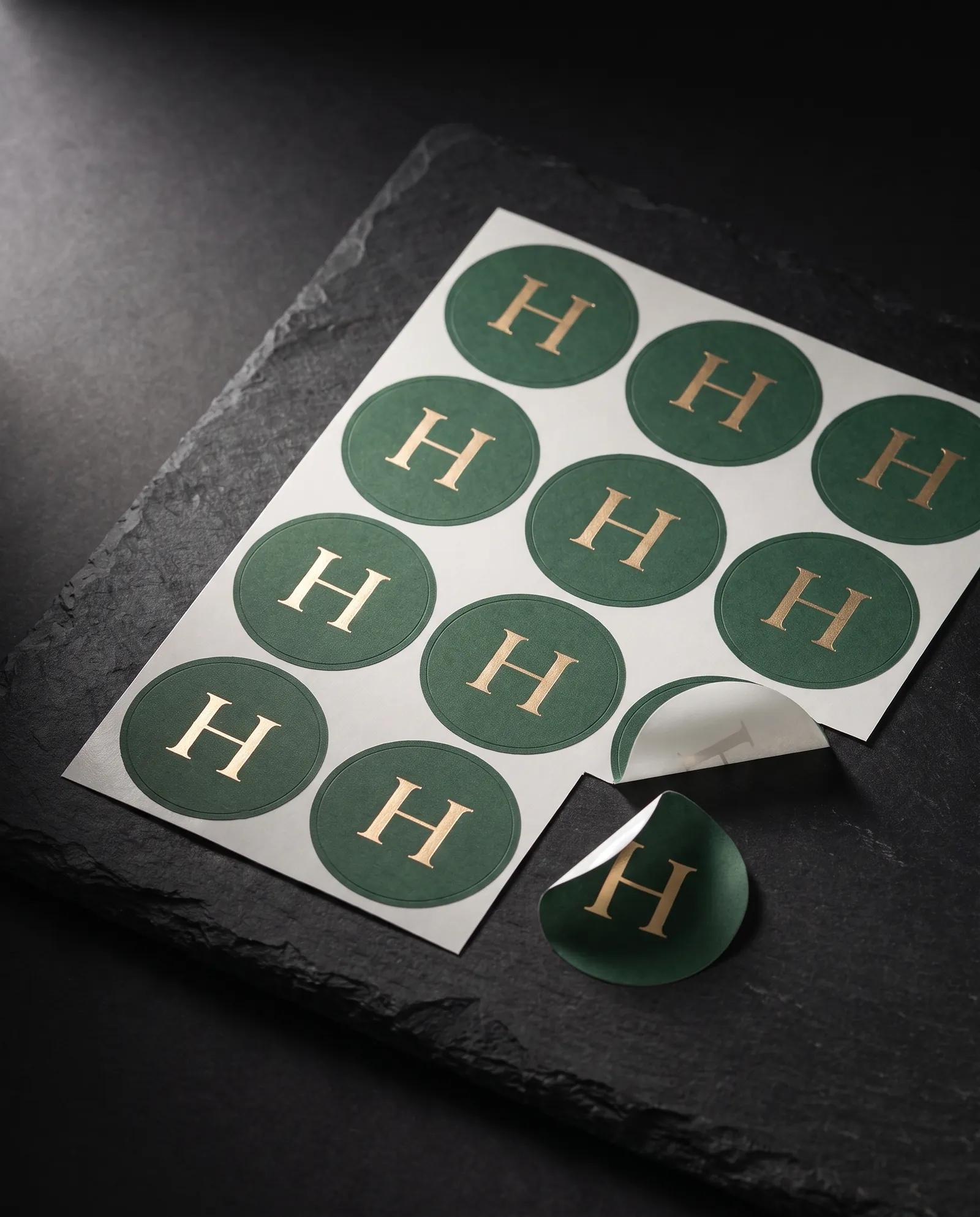 A kiss-cut sheet of round green stickers with a gold foil monogram, two peeled off