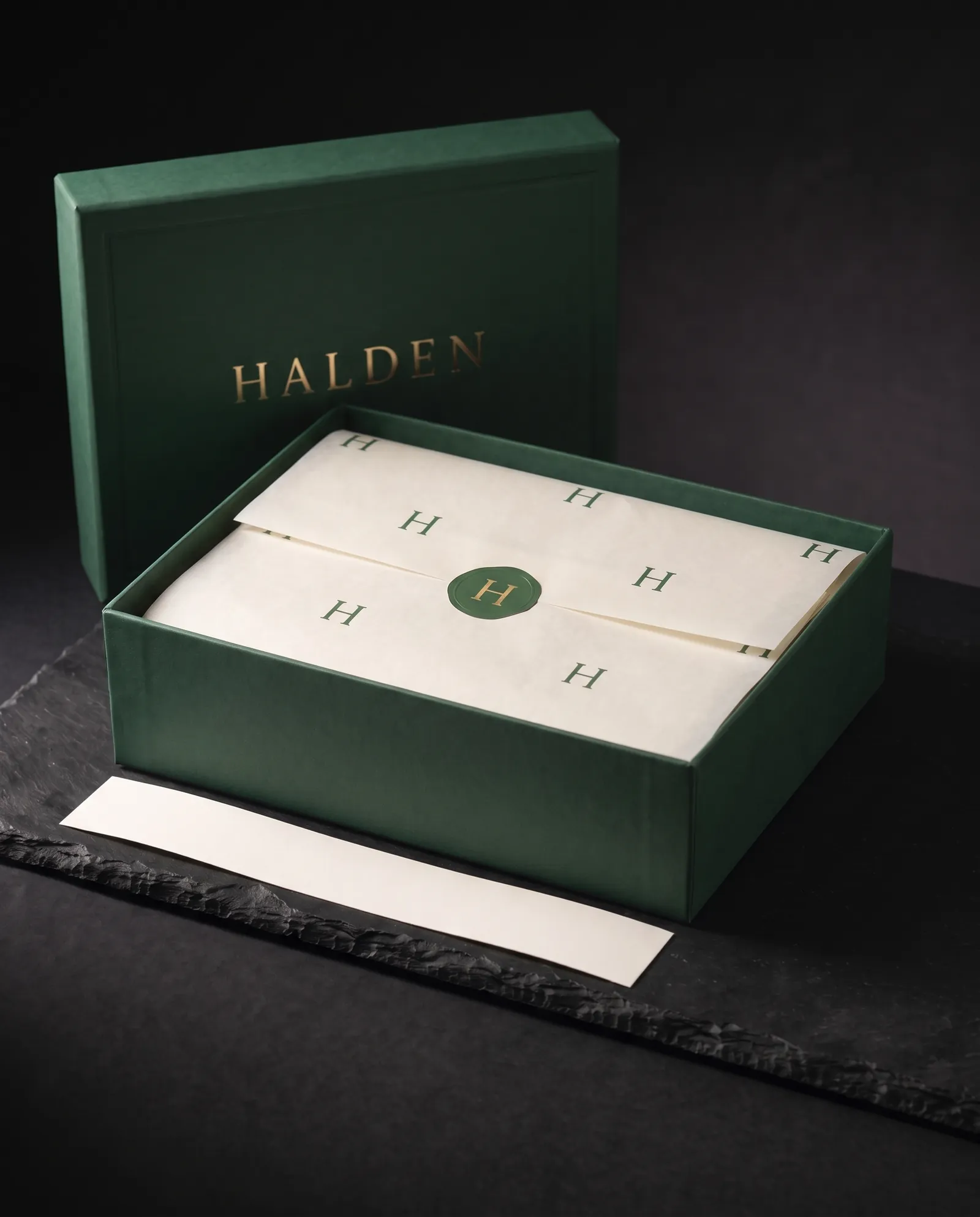 Lid lifted off the green box, printed tissue folded inside and closed with a round foil sticker seal