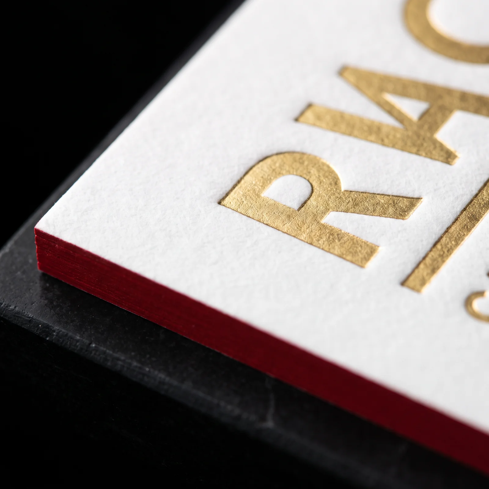 Close-up of gold hot foil pressed into cotton card stock with a deep red painted edge
