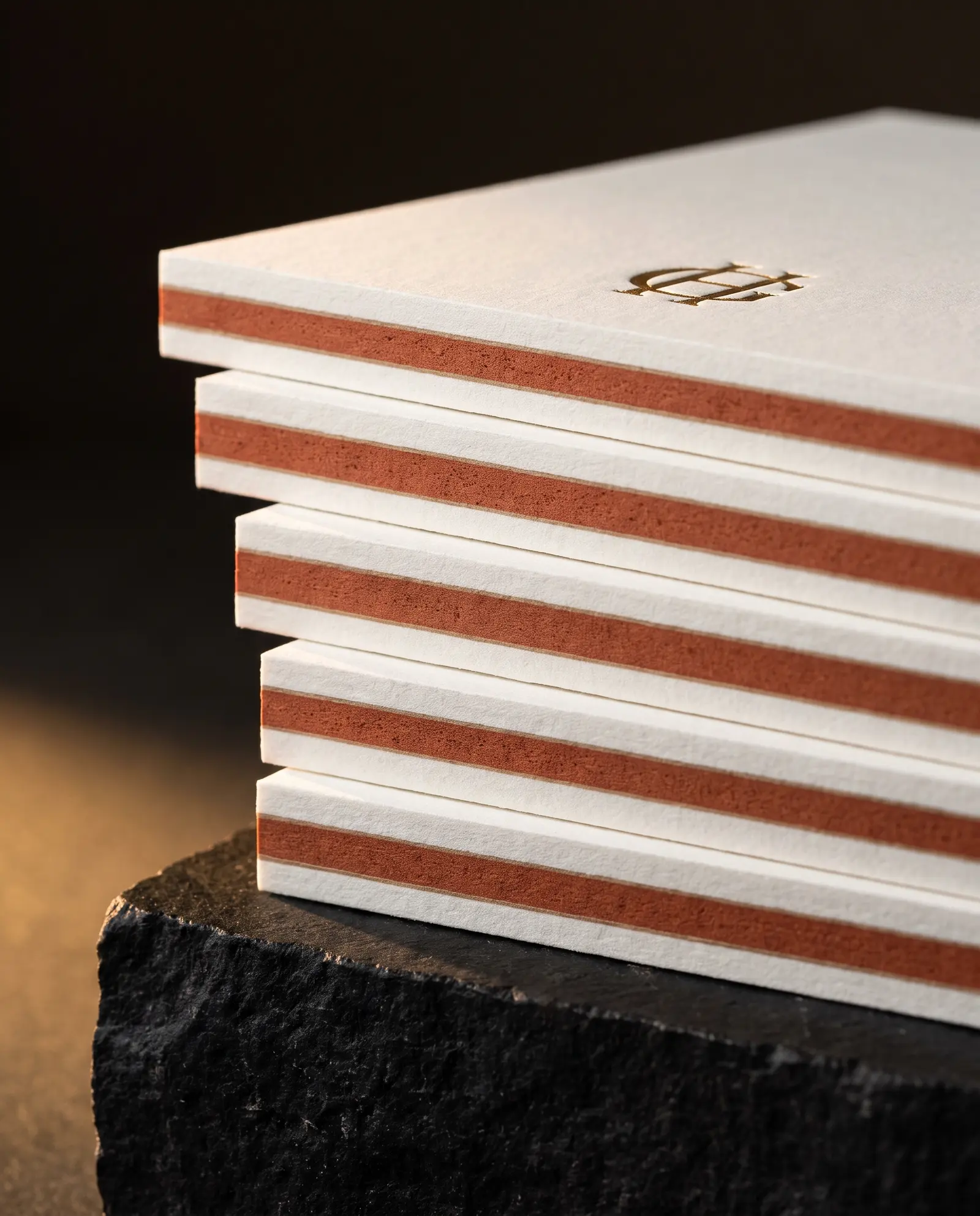 Edge of a stack of insert cards showing a terracotta core stripe between two white cotton sheets, the top card stamped with a gold foil mark.