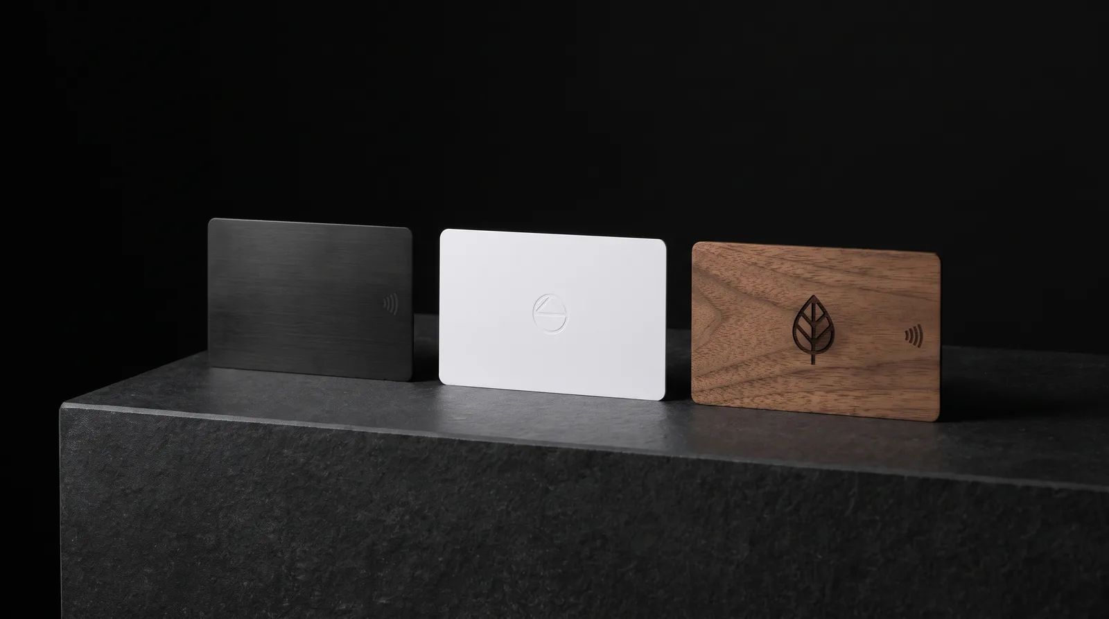 Three NFC cards on a black plinth: brushed stainless steel, white PVC and walnut wood