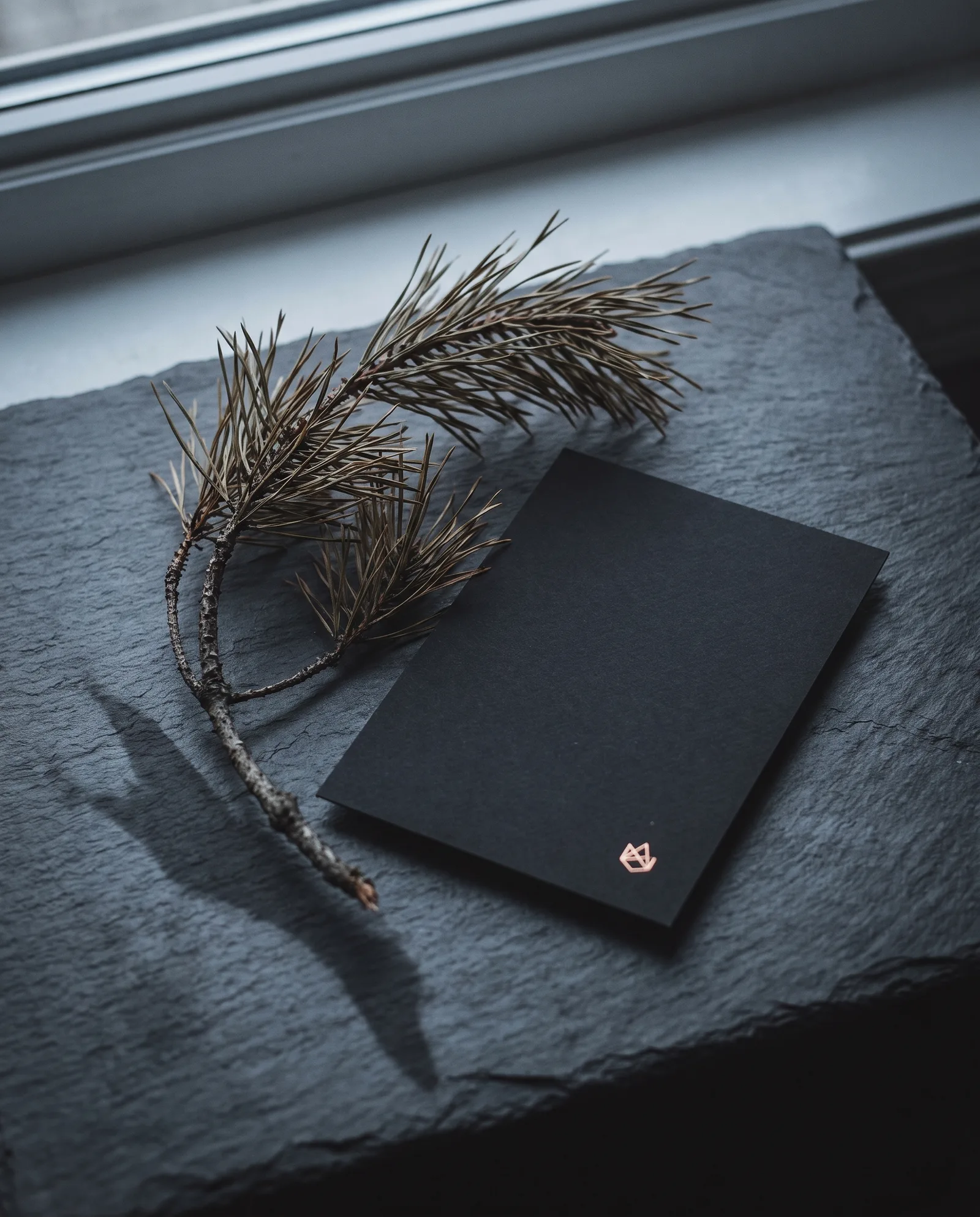 Black cotton holiday card with a copper hot-foil mark beside a dried pine sprig on dark stone