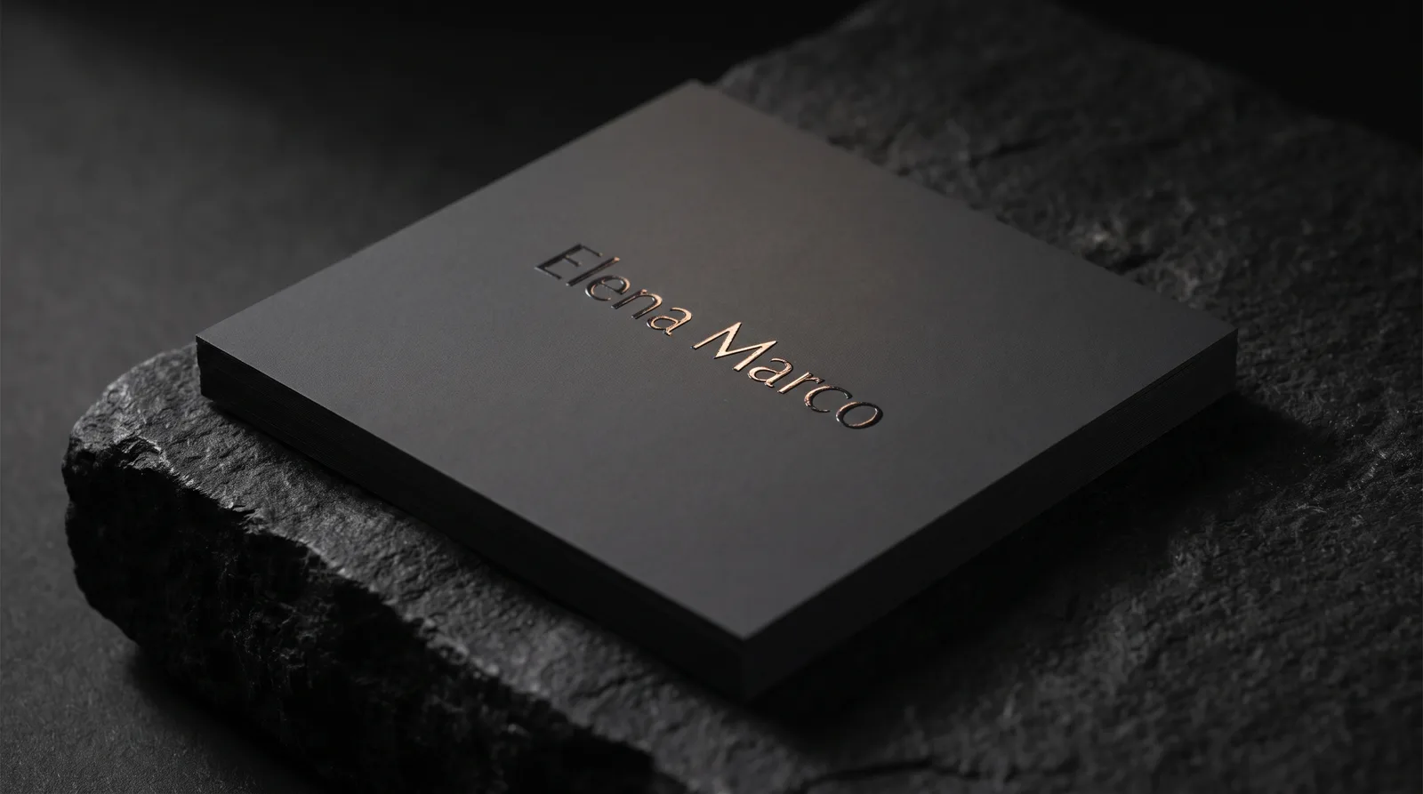 Charcoal soft-touch business card with raised clear spot UV wordmark catching warm light on black stone