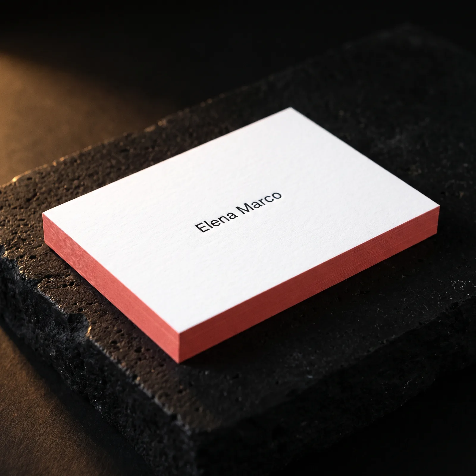 White business card with black name type and coral painted edge on dark stone