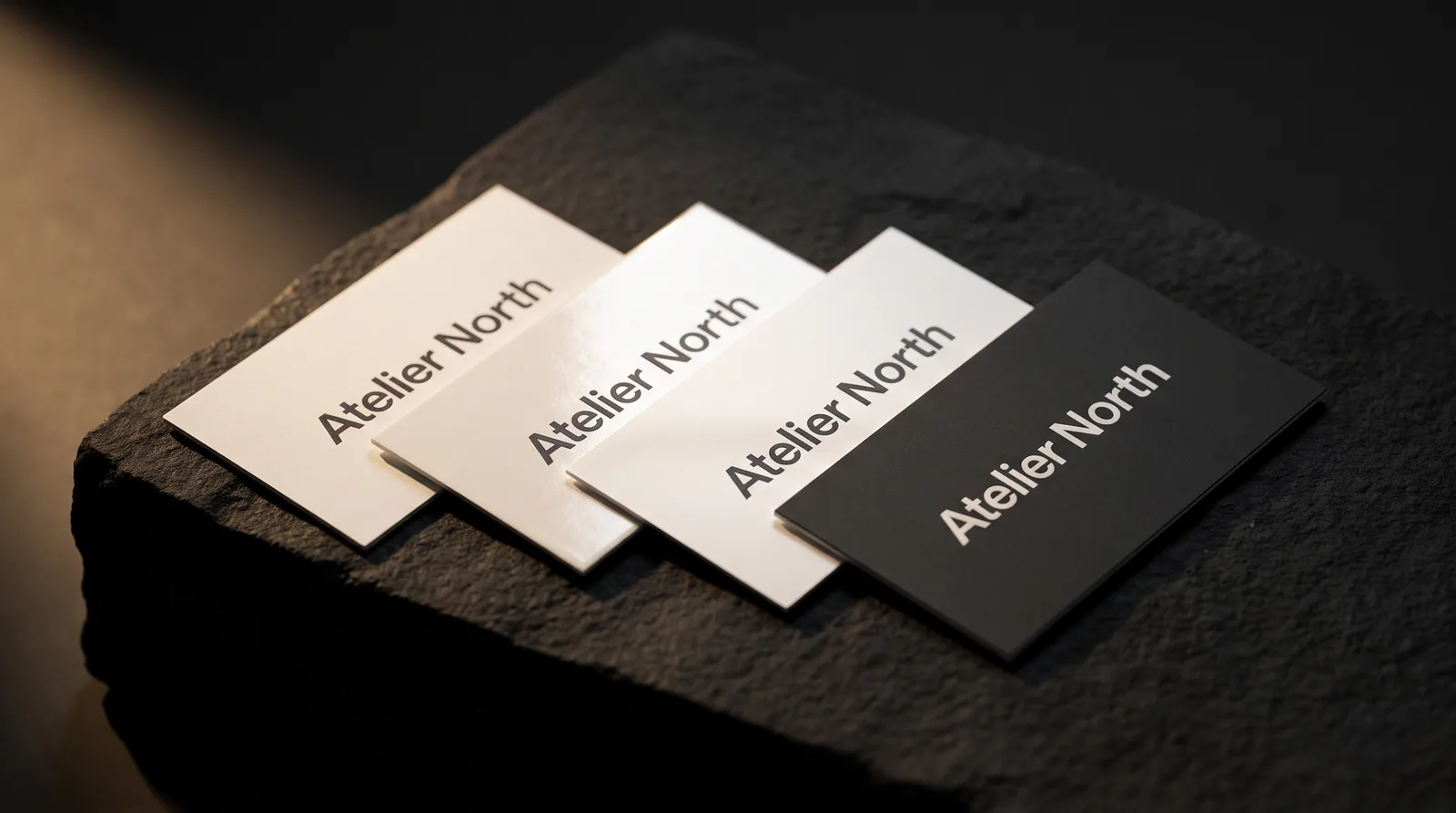 Four classic business cards on black stone: matte, glossy, silk and charcoal soft-touch under raking light