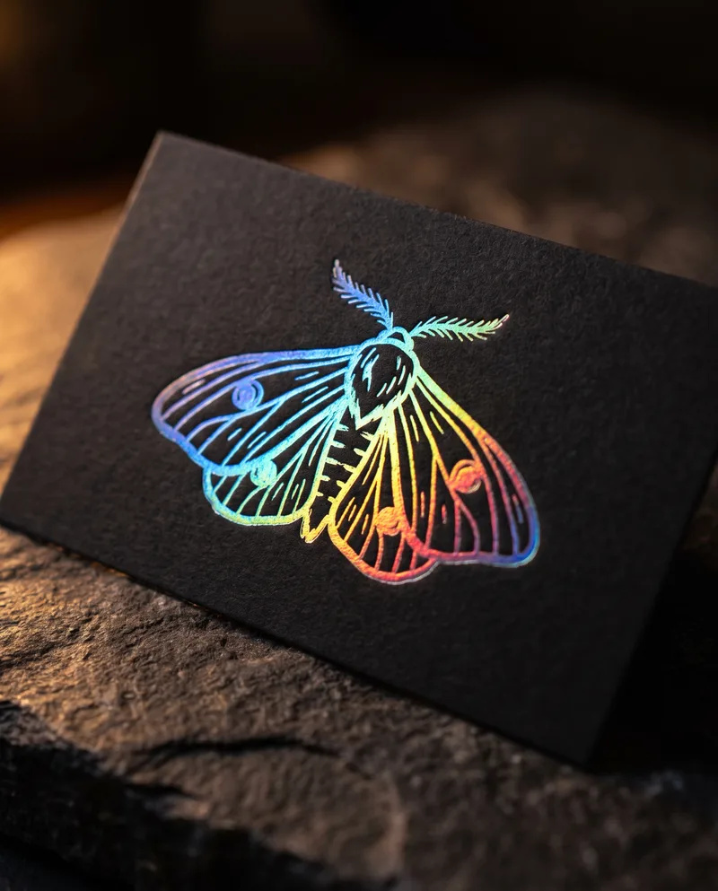 Macro of holographic foil moth illustration pressed into matte black business card under warm raking light
