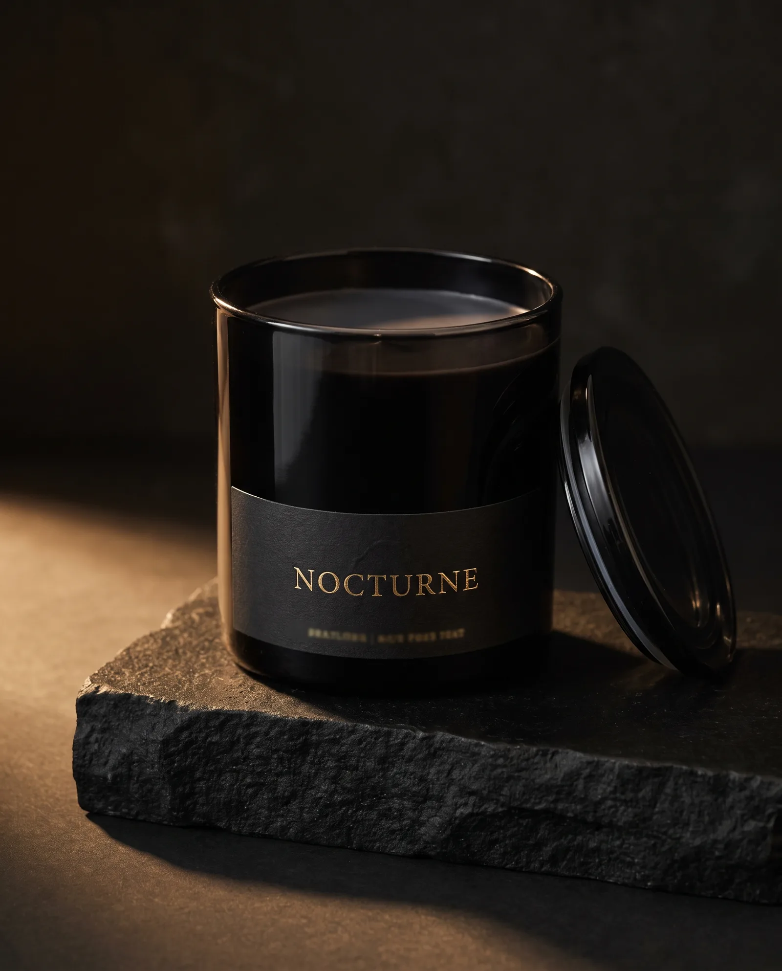 Black glass candle vessel with a black uncoated label carrying a gold foil wordmark, on dark stone.