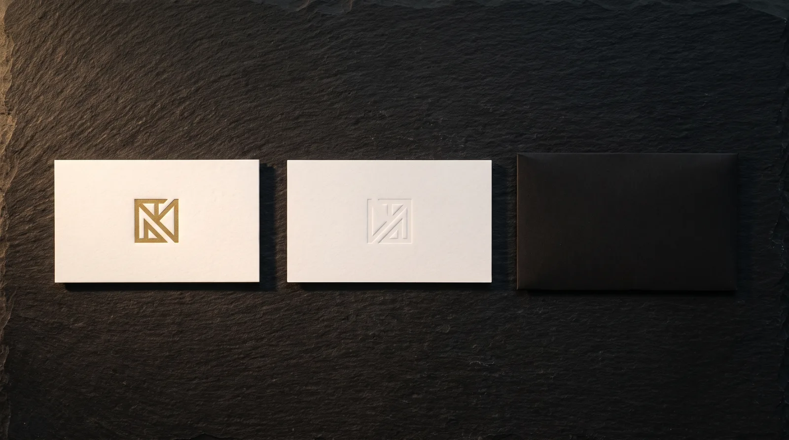 Gold foil monogram pressed into a deboss on a white cotton business card, with the back of a second card and a black sleeve