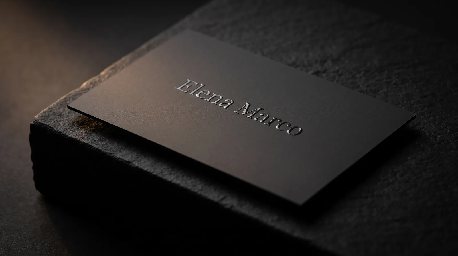 Raised spot UV gloss wordmark on a charcoal soft-touch business card catching warm raking light on black stone