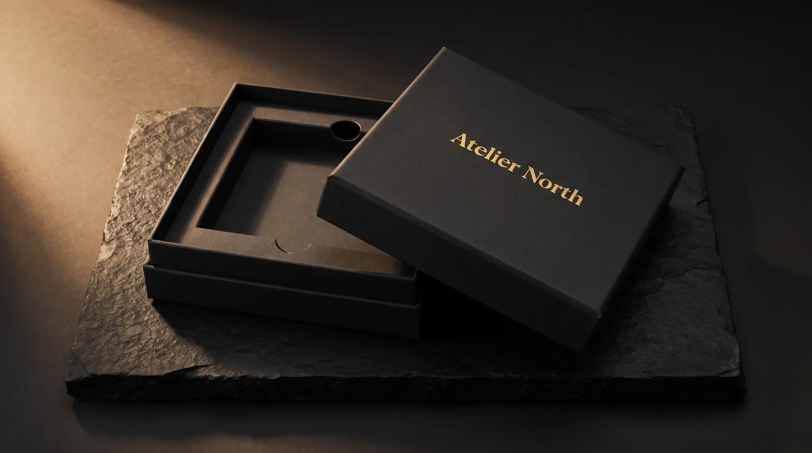 Black uncoated two-piece rigid box on black stone, gold hot-foil wordmark lid, paperboard tray inside