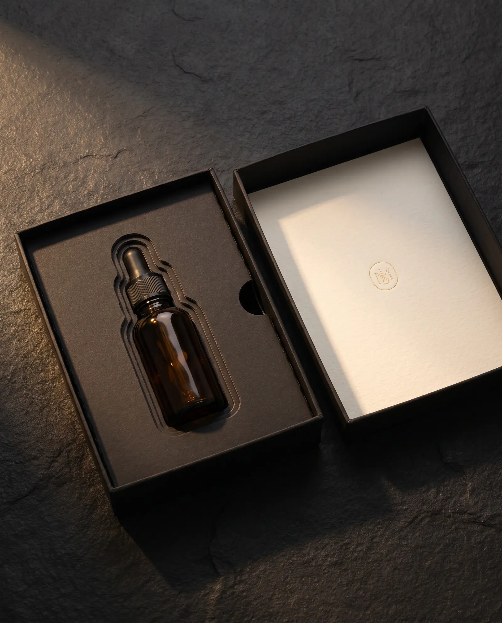 Top-down view of an open black box with a paperboard tray, a glass bottle seated in a cut recess beside a finger cut-out, and a cotton insert card in the lid.