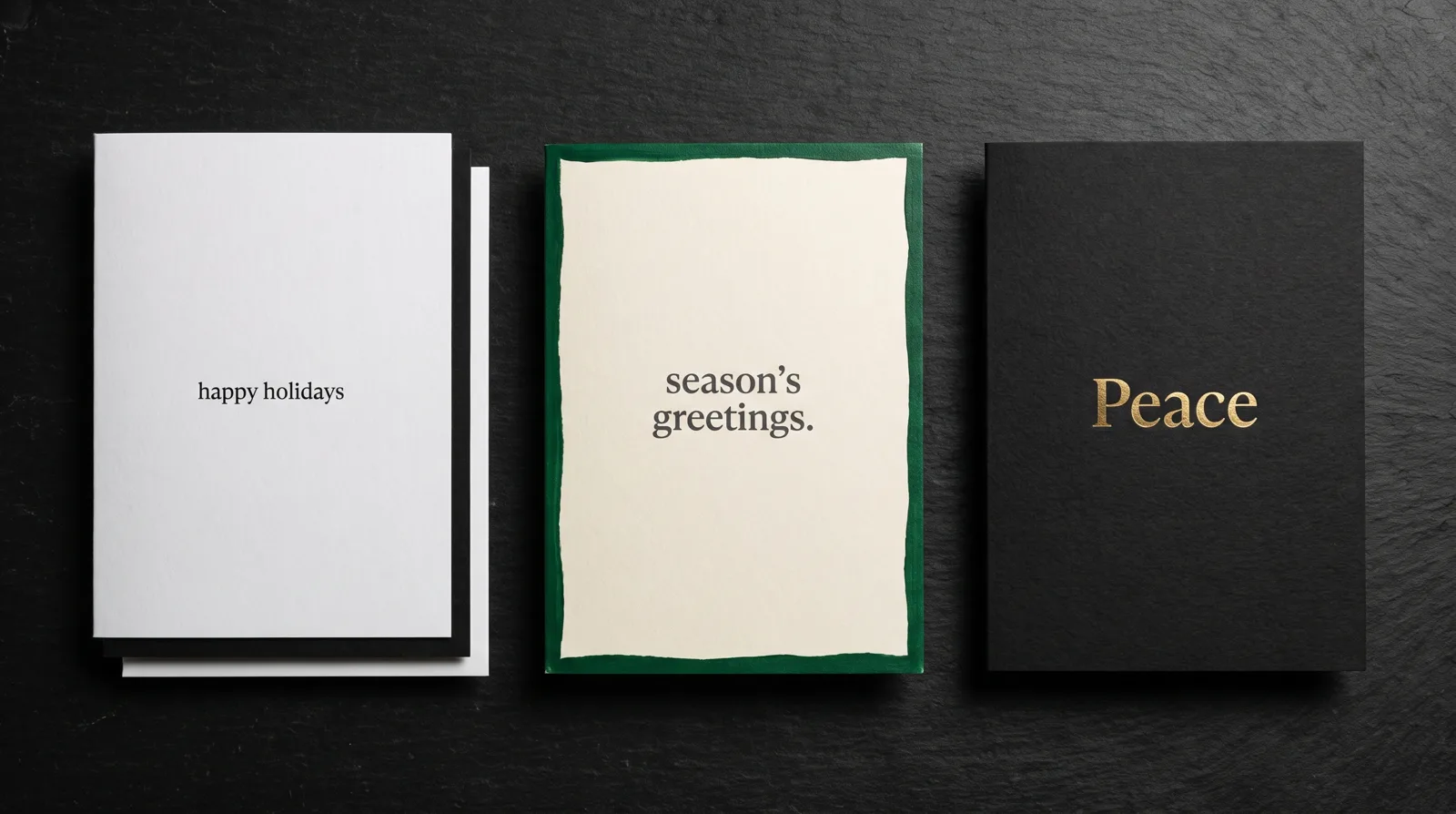 Three holiday cards side by side on black stone: a white card with a black core edge, a card with a forest-green painted edge, and a black card with Peace in gold foil.