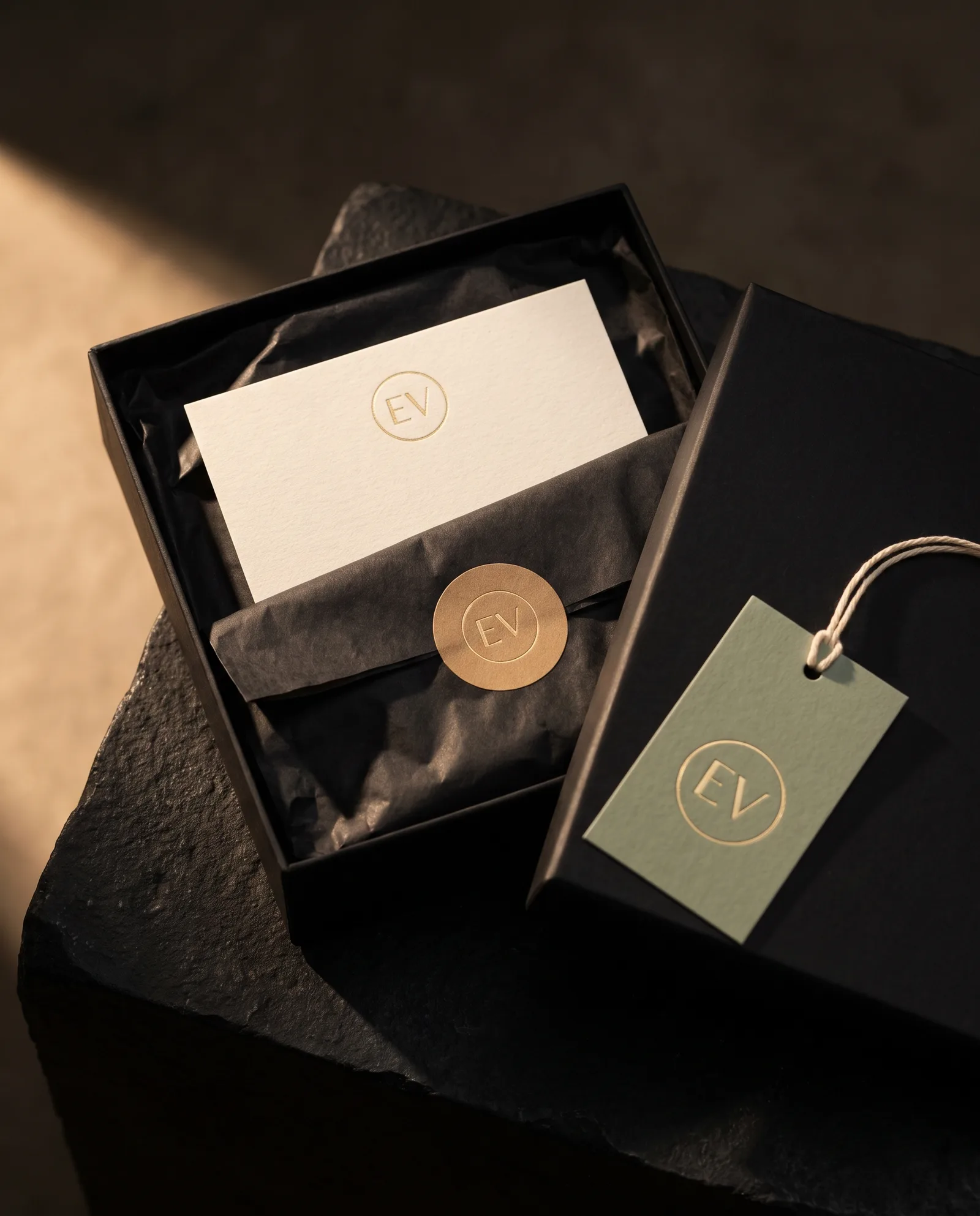 Black gift box open on dark stone, a cotton insert card with gold foil inside, a sage cotton gift tag on cotton string resting on the lid.