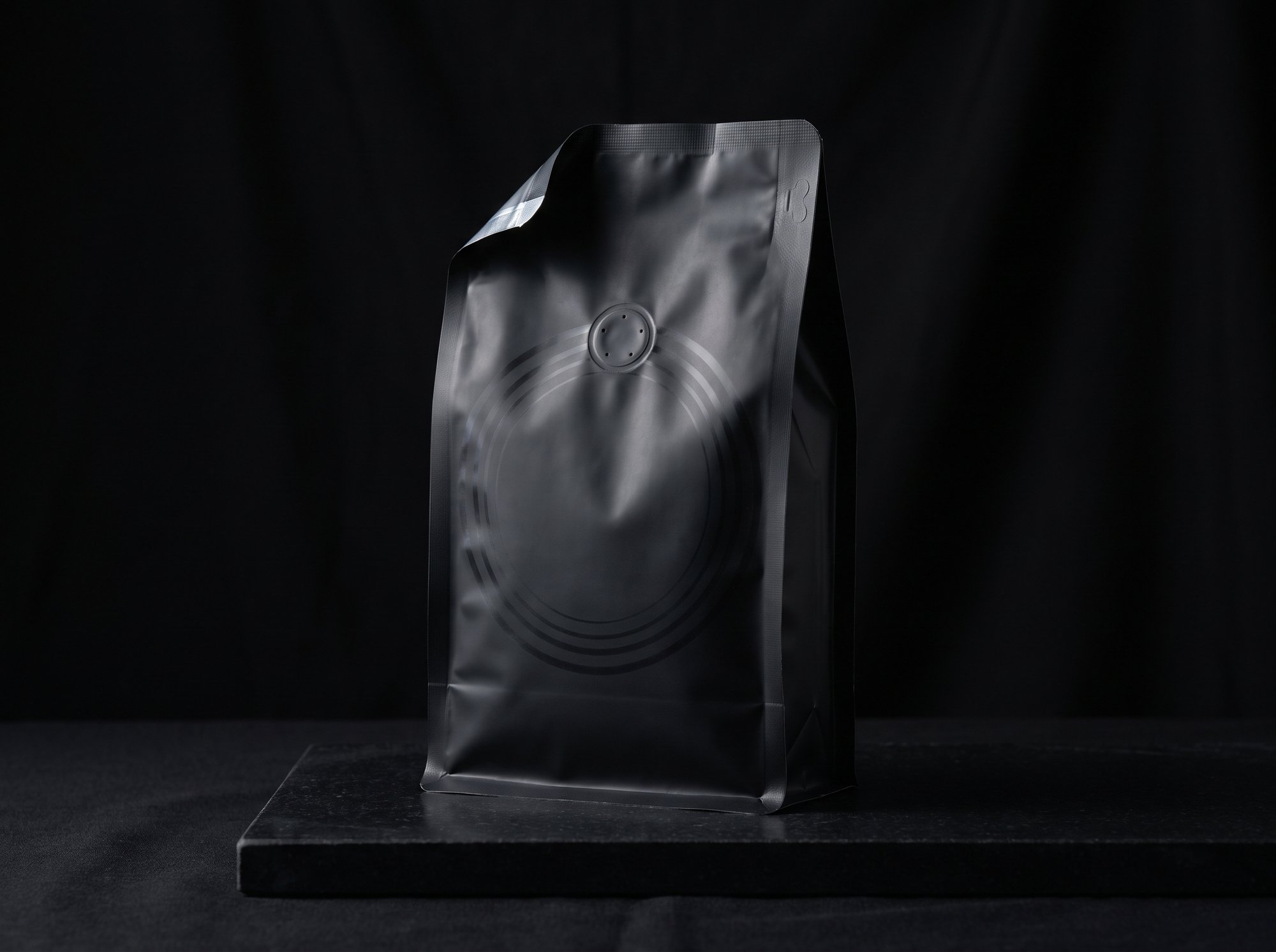 A soft-touch matte black coffee pouch with glossy varnish rings catching the light.
