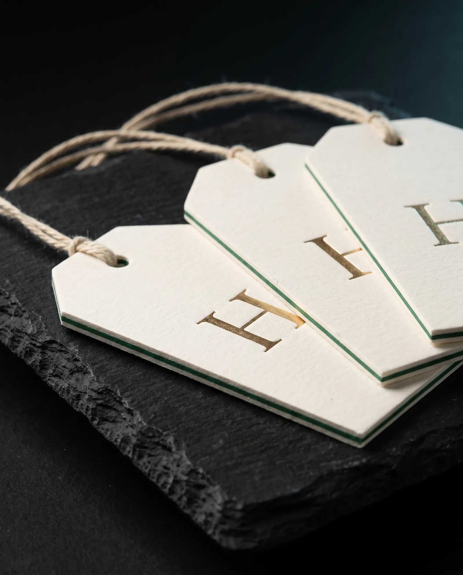 Three thick cotton hang tags on cotton string, gold foil monogram, a green duplex layer showing on the edge