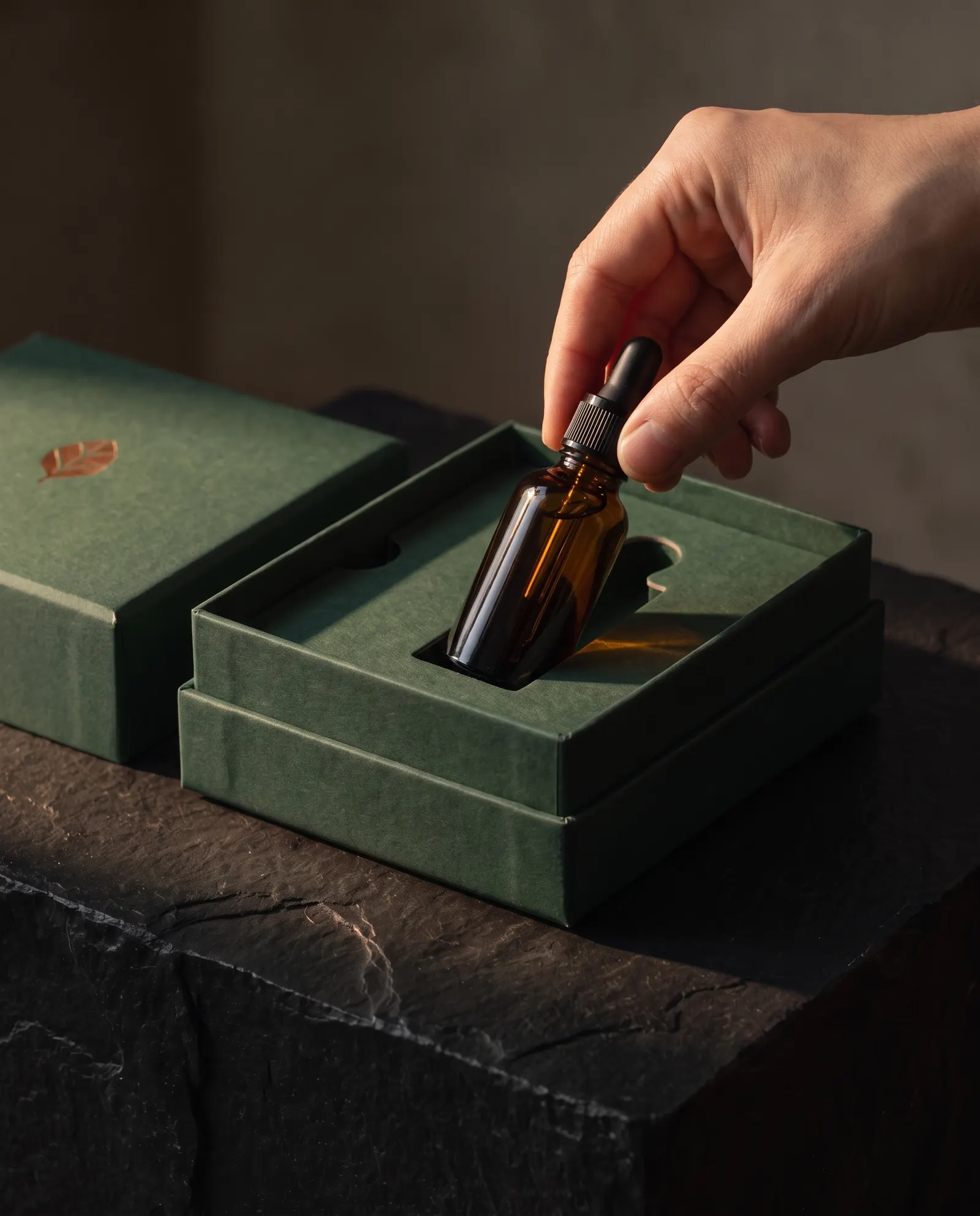 Hand lifting an amber bottle from a green rigid box's paperboard tray recess, no foam