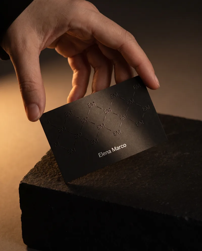 Hand tilting black soft-touch business card, repeating monogram in raised clear gloss, white foil name