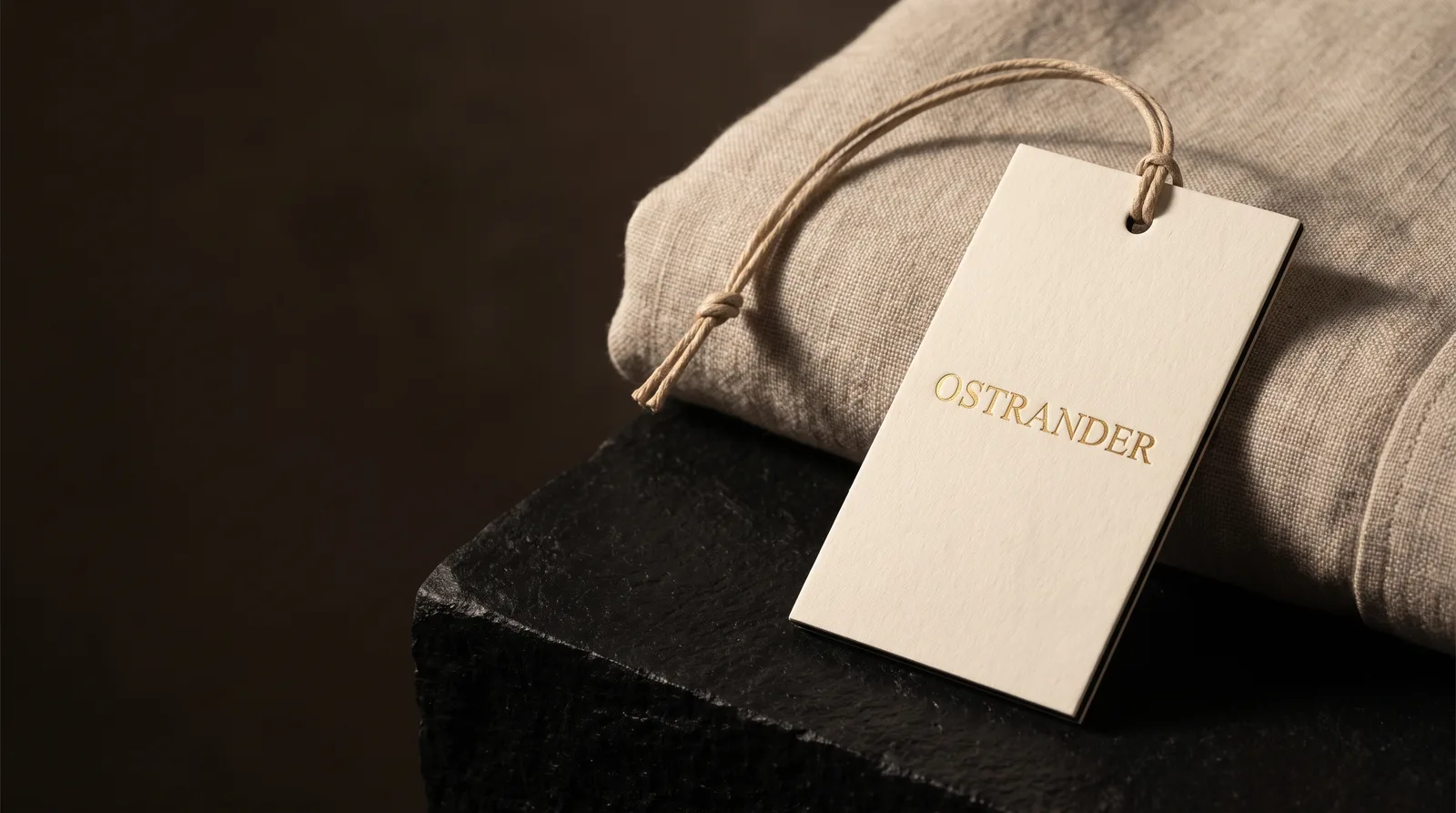 Thick cream cotton hang tag with a gold foil wordmark and a dark core stripe on its edge, hanging on waxed cotton cord from a folded linen garment.