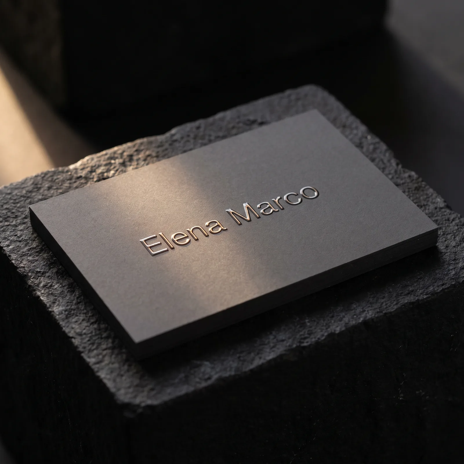 Charcoal soft-touch business card with raised clear gloss Elena Marco wordmark on stone plinth