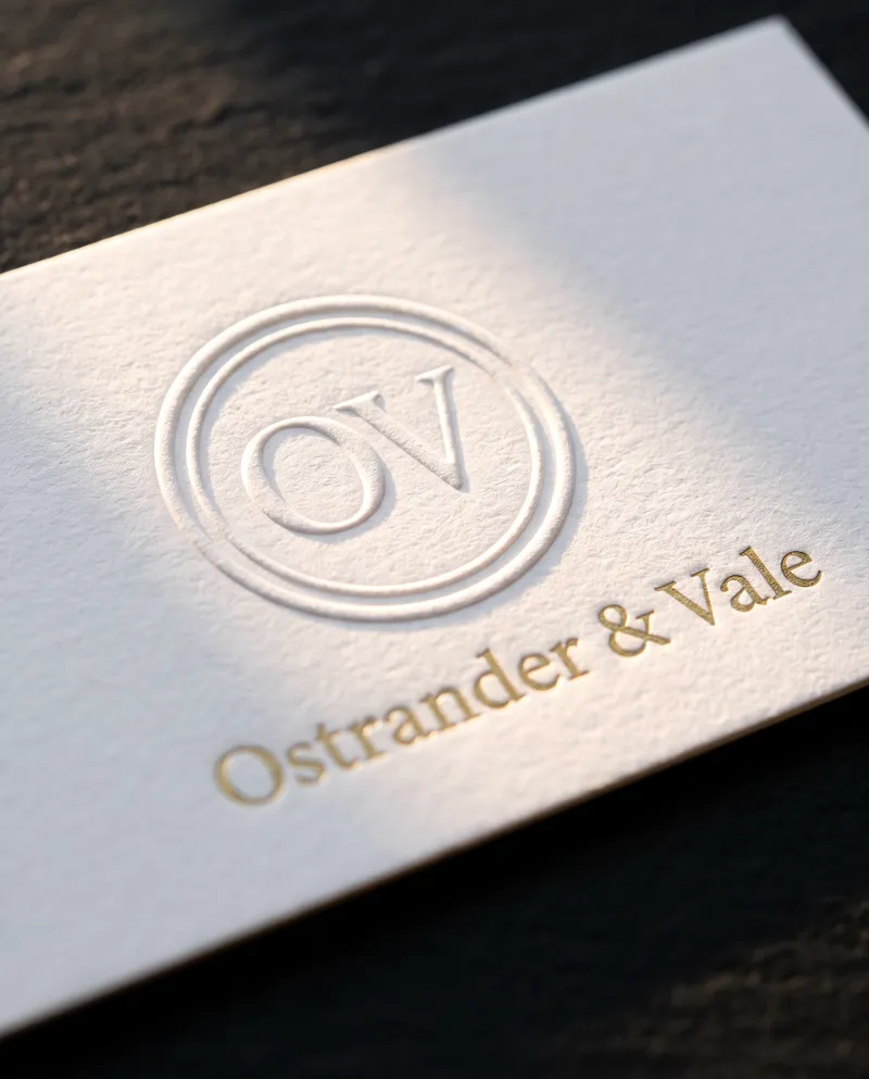 Macro of blind embossed OV monogram ring on white cotton card with gold foil name below