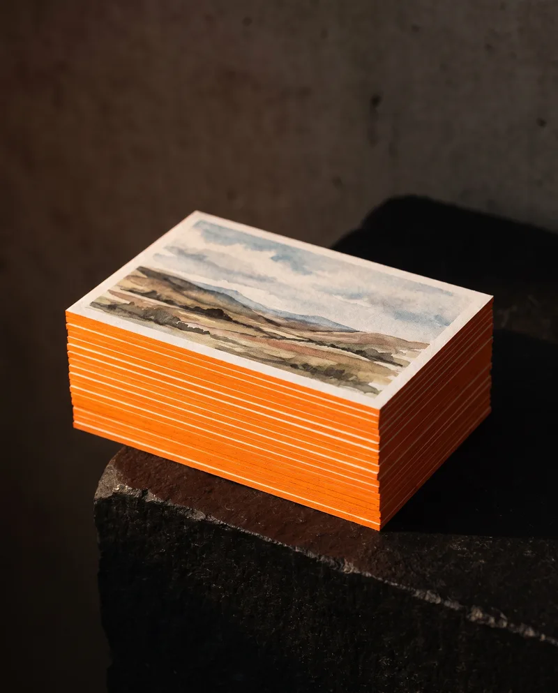 Stack of thick uncoated business cards with bright orange painted edges and printed watercolour landscape