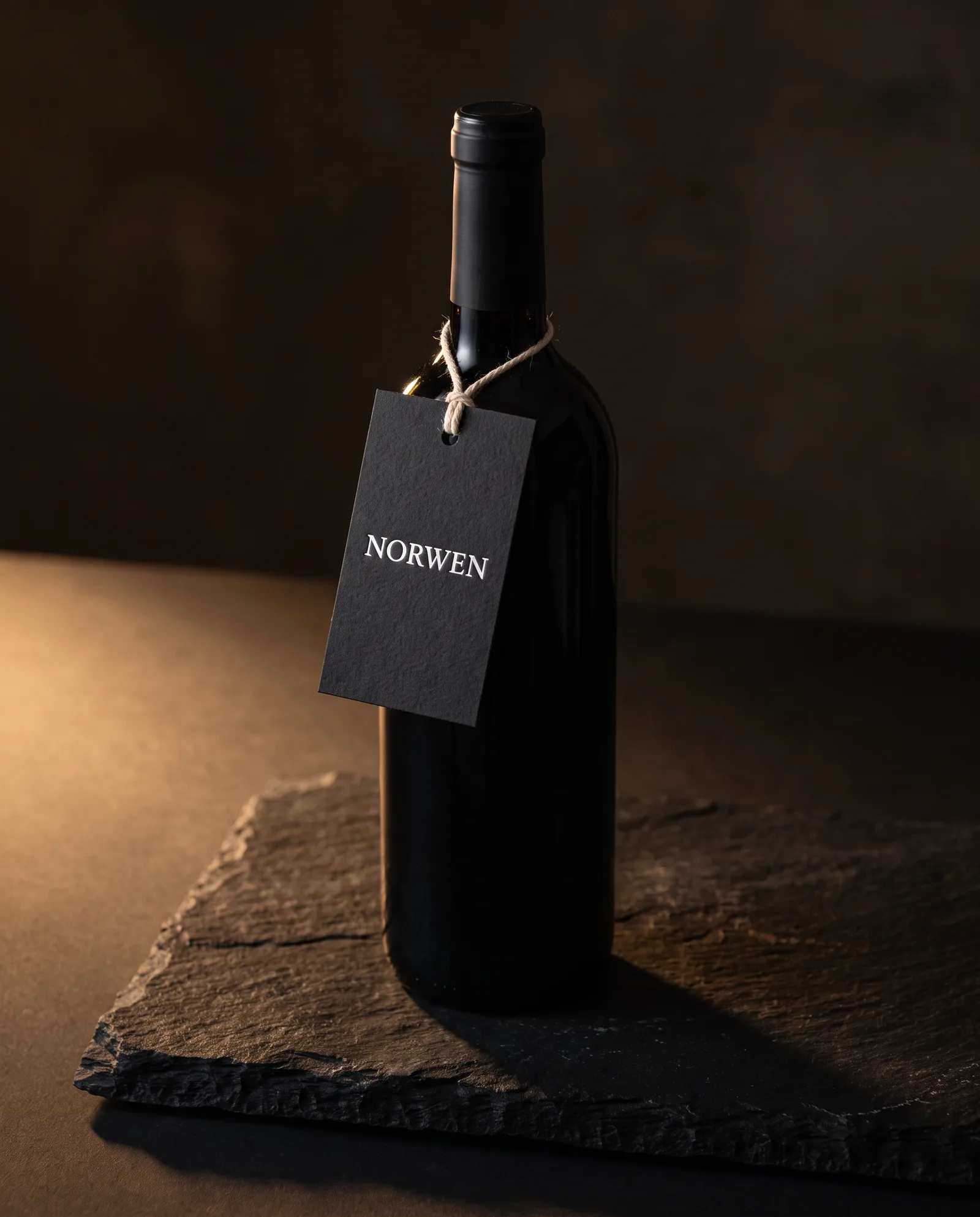 Dark wine bottle on black stone with a black cotton tag in white pigment foil tied at the neck with natural cotton string.